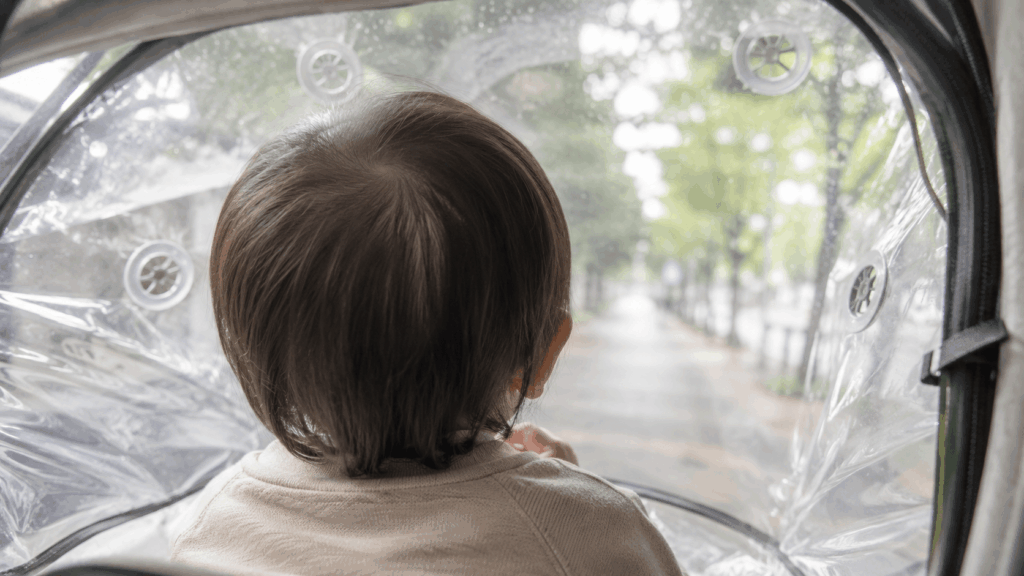 透明レインカバー越しにベビーカーから外を眺める幼児の後ろ姿