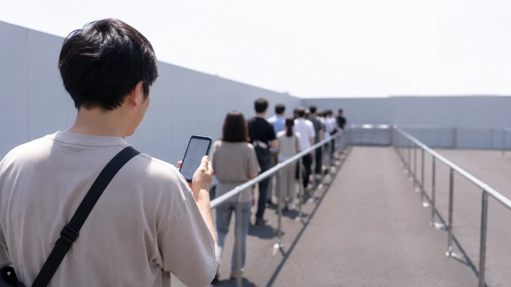 屋外の列に並びながらスマートフォンで待ち時間を確かめている人の後ろ姿