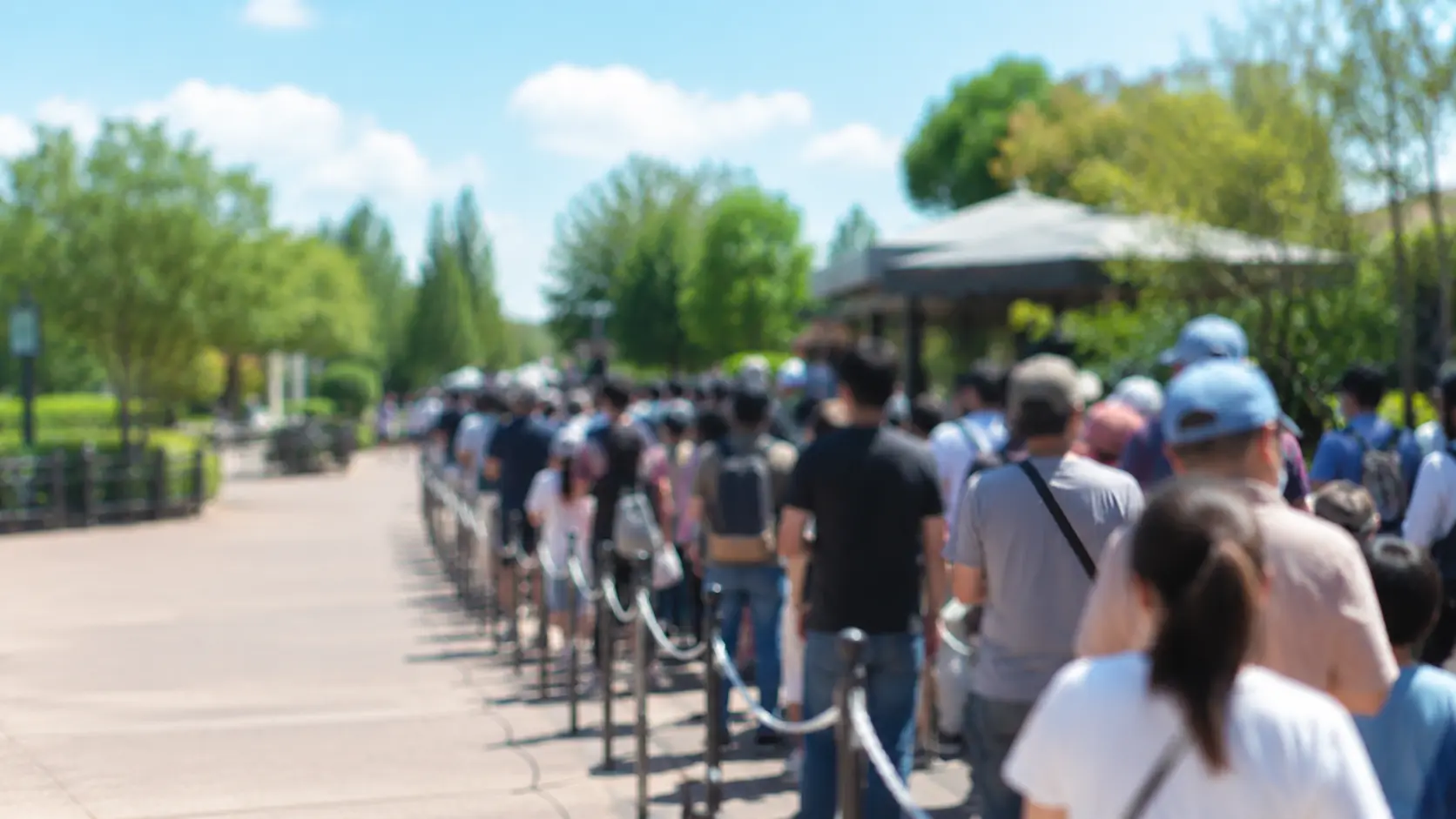 夏の遊園地で柵とロープに沿って待機列に並ぶ人々を遠景から撮影した様子